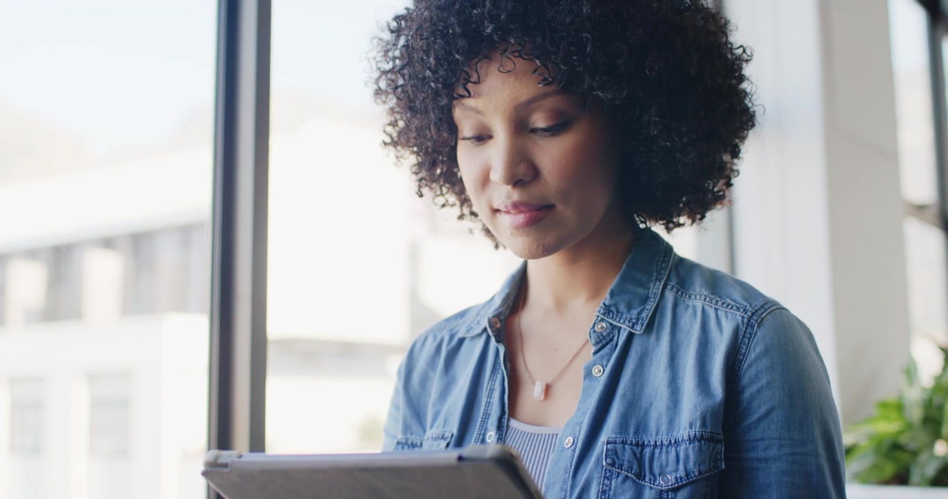 African American woman holding tablet, tapping, scrolling while reading at window with smartwatch