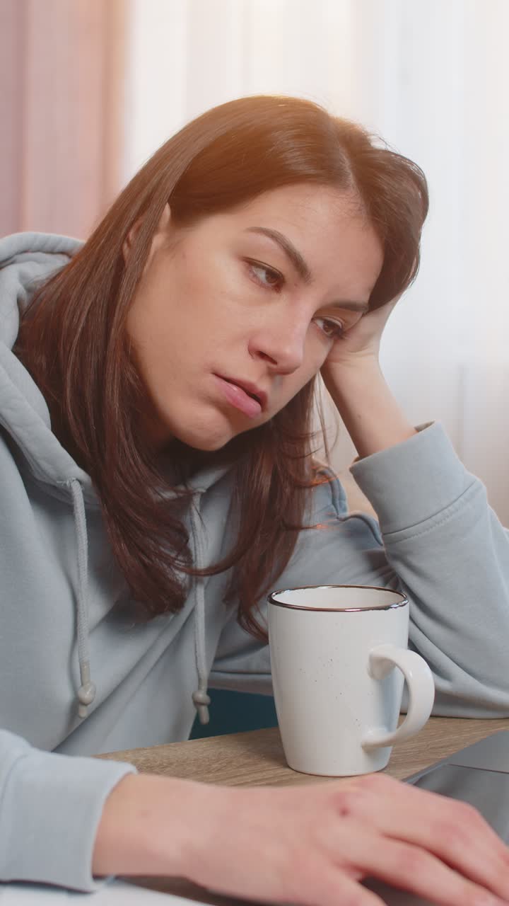 Tired young woman freelancer using laptop computer at table with documents in living room at home