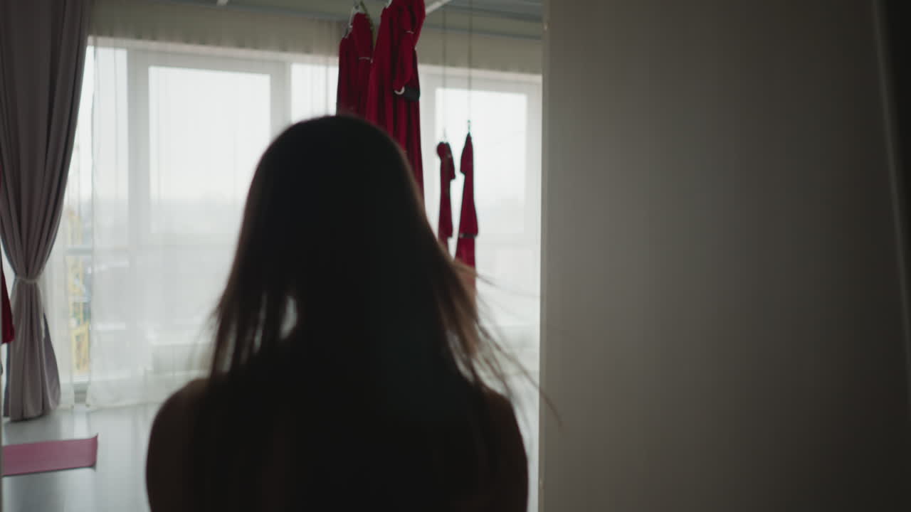 Back view yoga instructor walking into airy studio with suspended red yoga hammock hanging by window with sheer curtains under soft natural light capturing serene practice environment