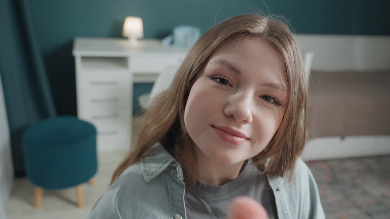 Ragazza adolescente bianca che applica il pennello da trucco in camera da letto, primo piano, influencer giocosa che prova il fard e posa per la fotocamera, luce soffusa del mattino, arredamento accogliente, lampada e sgabello visibili, sorriso naturale e sicura di sé.