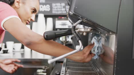 happy biracial female barista weearing apron cleaning coffee machine in cafe