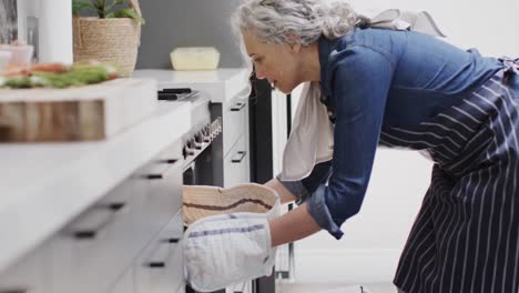 happy senior caucasian woman taking out baked vegetables from oven in kitchen, slow motion