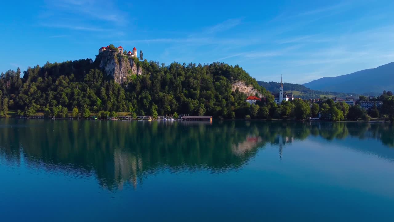Aerial 4K drone video over Lake Bled on a sunny summer morning overlooking Bled Castle and Bled Town with stunning reflections in the clear water