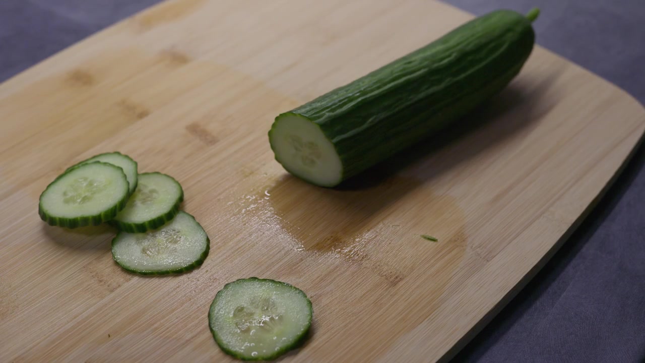 Primo piano di mani con guanti che affettano un cetriolo fresco con un coltello da cucina su un tagliere di legno durante la preparazione del cibo