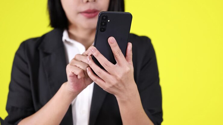 Donna con blazer nero che usa uno smartphone, mezza figura, sfondo giallo vivace, illuminazione uniforme da studio