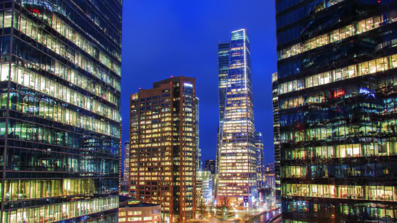 A stunning view of a vibrant city skyline at night showcases tall buildings with glowing windows and reflective glass facades. The atmosphere captures the bustling energy of urban life.