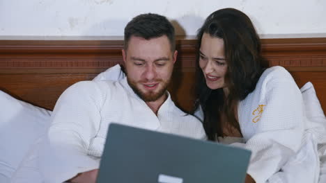 young couple spend time together. beautiful woman and her husband in white dressing gowns laying on bed and looking into the laptop.