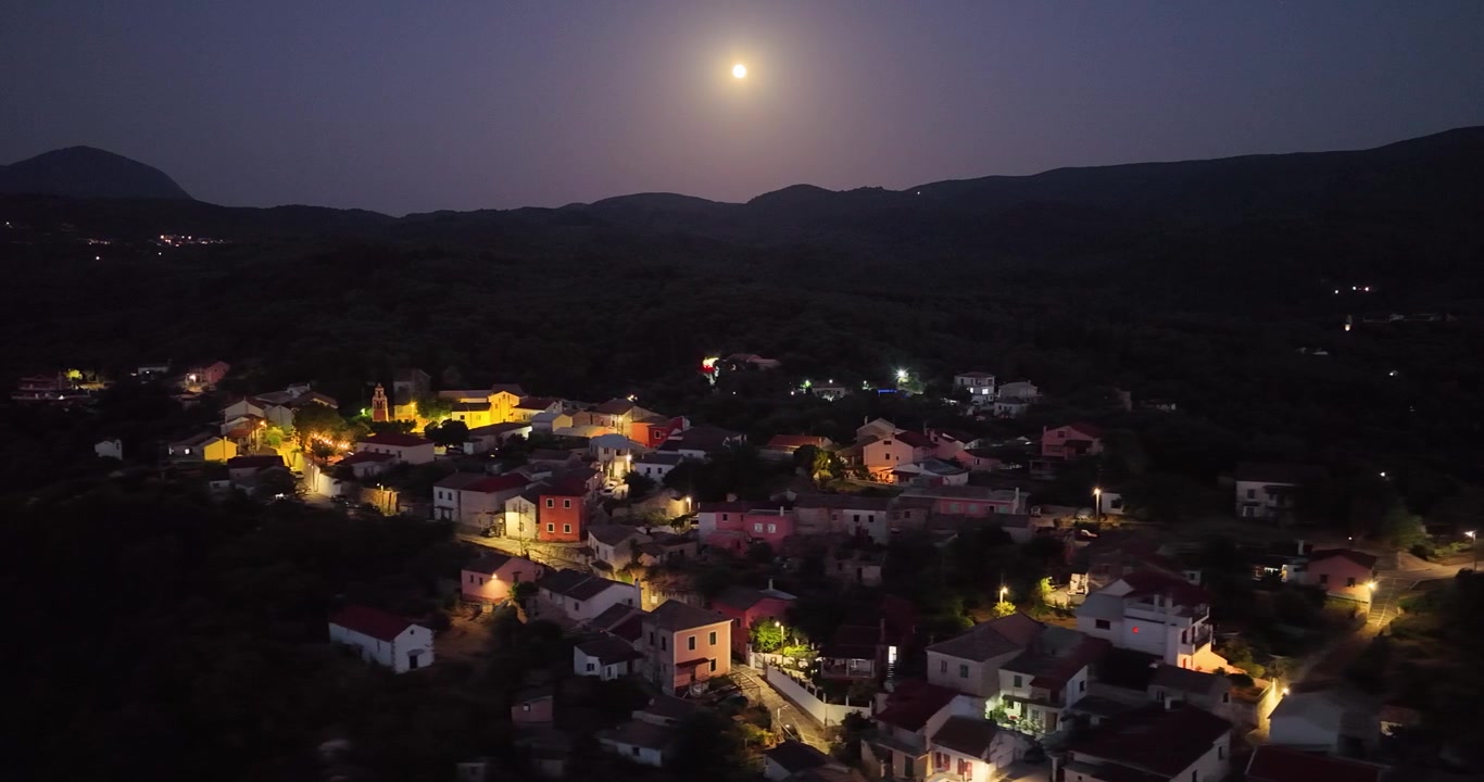 Vista panorâmica aérea de drone da vila de Agioi Douloi na ilha de Corfu, na Grécia, à noite, sob uma lua cheia brilhante, com casas iluminadas e colinas escuras