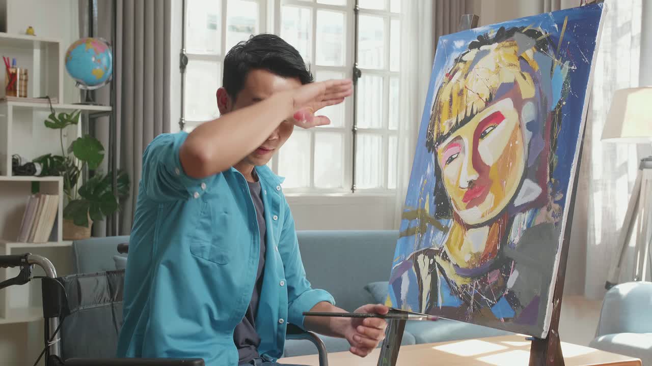 Side View Of An Asian Artist Man In Wheelchair Holding Paintbrush Turn To Wipe The Sweat Before Crossing His Arms And Smiling While Painting A Girl On The Canvas