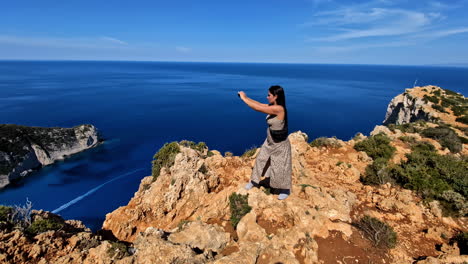una mujer caucásica se encuentra muy cerca en el borde rocoso de una colina empinada en navagio, grecia.