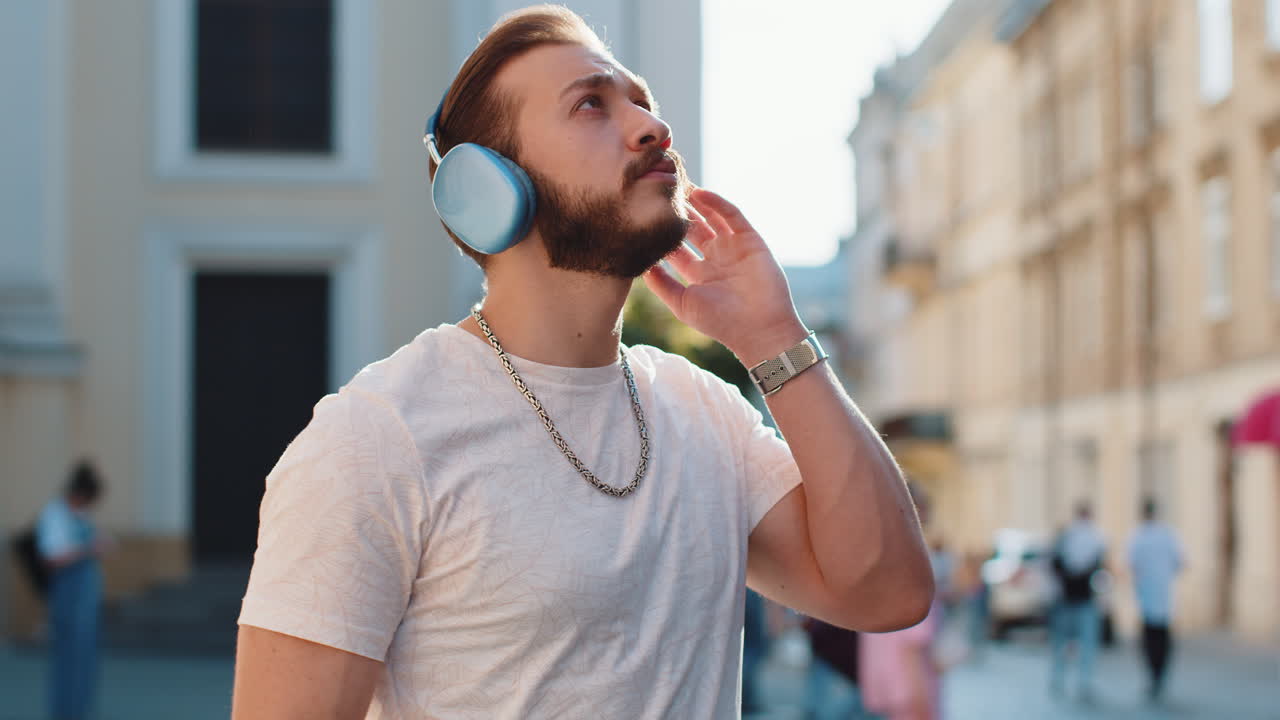 Happy young adult man in wireless headphones choosing listening music dancing outdoors city street