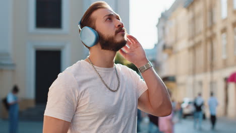 happy young adult man in wireless headphones choosing listening music dancing outdoors city street