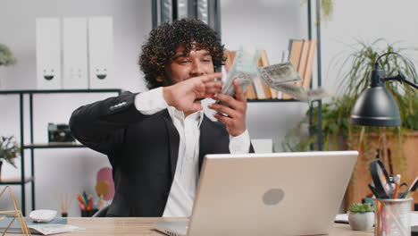 Cheerful-rich-business-man-working-on-office-laptop-wasting-throwing-money-to-camera-profit-win