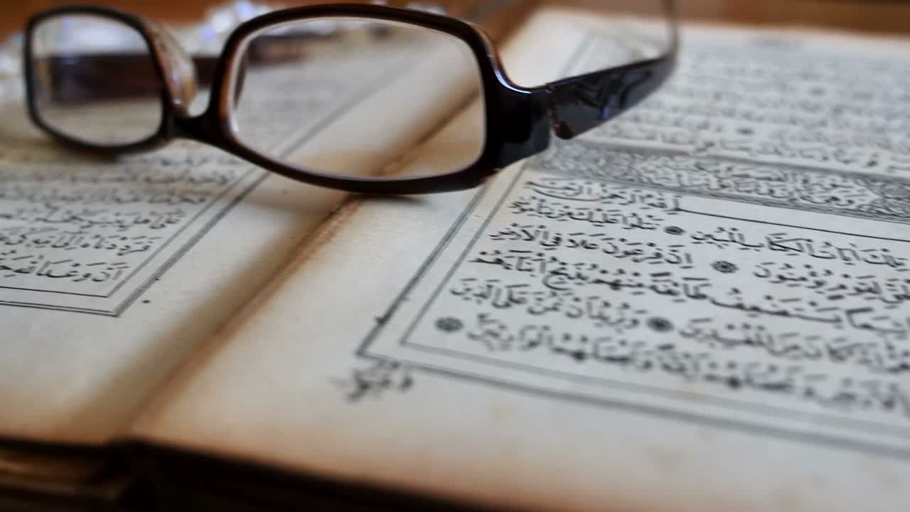 Footage of a Quran lying on a table with reading glasses on it and a tasbeeh in the back, the shot is moving from the left to the right