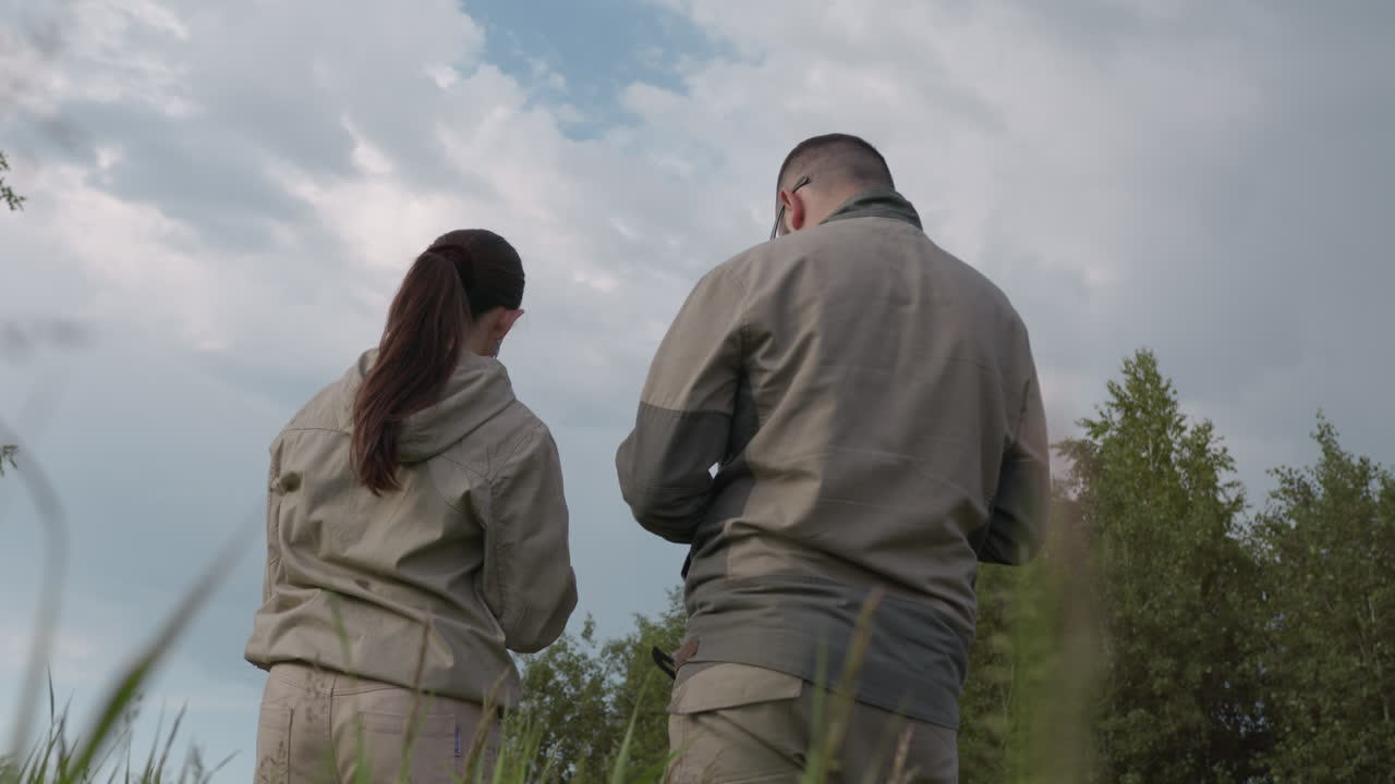 back view of two adults in open field conducting survey work, wearing khaki jackets and gloves, holding devices while observing sky and surroundings under cloudy sky, scattered trees and tall grass
