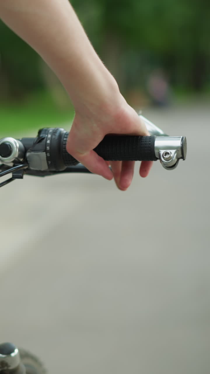 vista parcial de alguien con camiseta negra caminando de cerca con su bicicleta a lo largo de un camino tranquilo y pacífico, con el enfoque en su mano sosteniendo el manillar de la bicicleta, la carretera está rodeada de vegetación