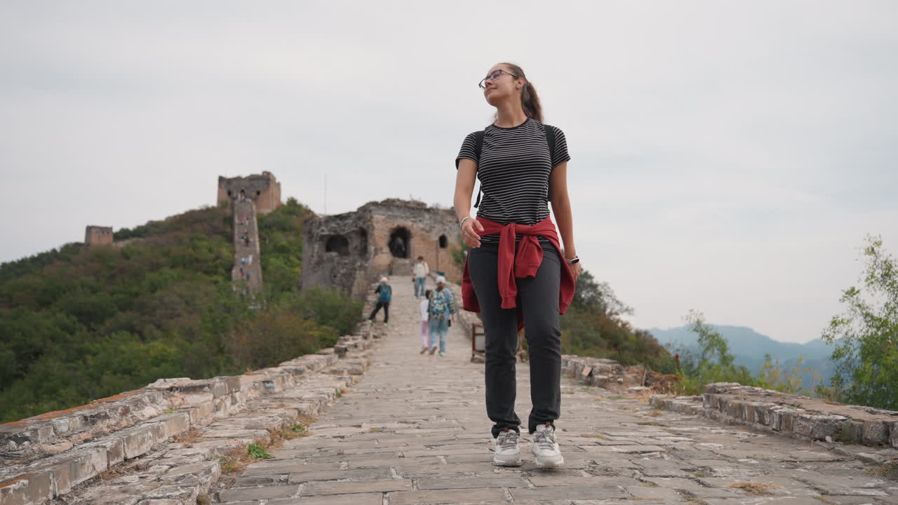 Donna bianca che esplora un affollato sentiero in rovina con un piccolo gruppo, occhiali da sole e un maglione rosso legato in vita, partecipante casuale al tour che osserva altri visitatori e il paesaggio, vivace scena turistica in un sito storico.