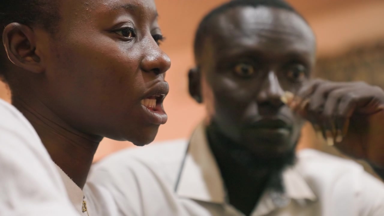 Pareja afroamericana compartiendo un momento íntimo y tierno. Sonrisas nerviosas en medio de una suave iluminación ambiental revelan vulnerabilidad. Pareja de estudiantes sentada en el interior intercambiando miradas sutiles con una tierna tensión emocional.