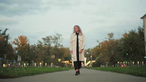 young woman in white coat holding umbrella walking along urban pathway glancing around with thoughtful expression surrounded by soft evening light, bokeh lights, blurred cars, trees, and structures