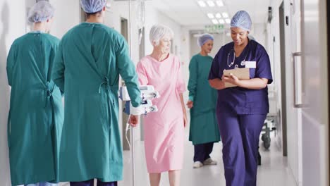 video of diverse female doctor and senior female patient on drip talking in busy hospital corridor