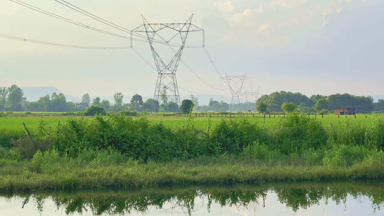High-voltage power transmission towers rise above lush farmlands beside a calm canal. Electric lines stretch across the open countryside under gentle daylight, symbolizing progress and connectivity