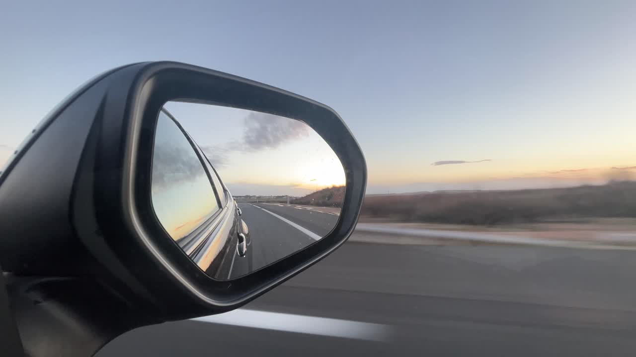 Un'auto sta percorrendo una strada e il conducente sta guardando nello specchietto retrovisore. La strada è vuota e il cielo è blu.