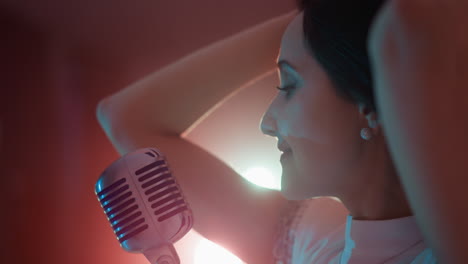 색 드레스를 입은 여성 가수가 빈티지 마이크 앞에 서서 머리에 손을 고 미소 짓고 머리를 부드럽게 움직이고 있습니다. 배경에는 조명이 있습니다.