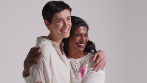 Close-Up-Studio-Portrait-Of-Two-Smiling-Women-Wearing-Pink-Breast-Cancer-Awareness-Ribbons-Hugging-Against-White-Background