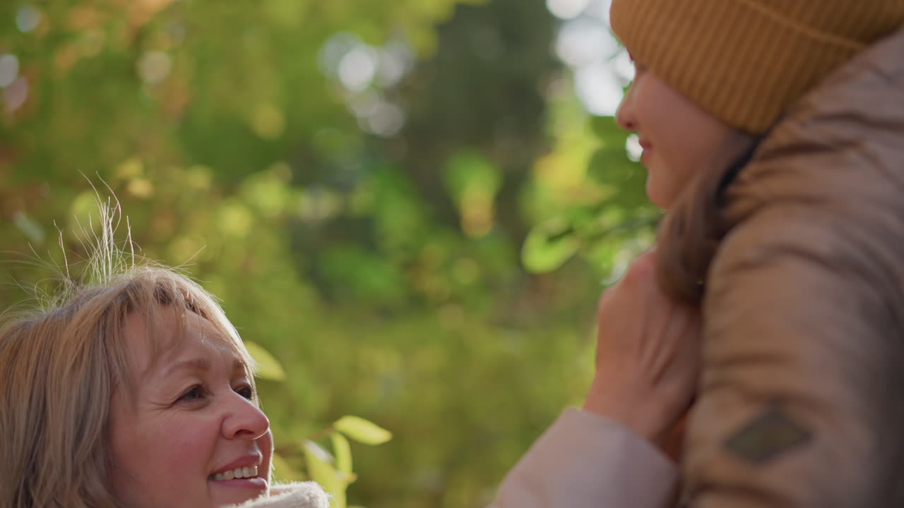 caring mother gently adjusts daughter hoodie in sunlit autumn woods as warm sunlight filters through colourful foliage, both dressed in jackets and hat