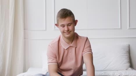 Niño-Con-Síndrome-De-Down-Sonriendo-A-La-Cámara-Sentado-En-La-Cama-En-El-Dormitorio-En-Casa