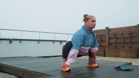 Sporty-fit-woman-in-blue-pink-sportswear-doing-squats-yoga-stretching-exercising-on-roof-of-house