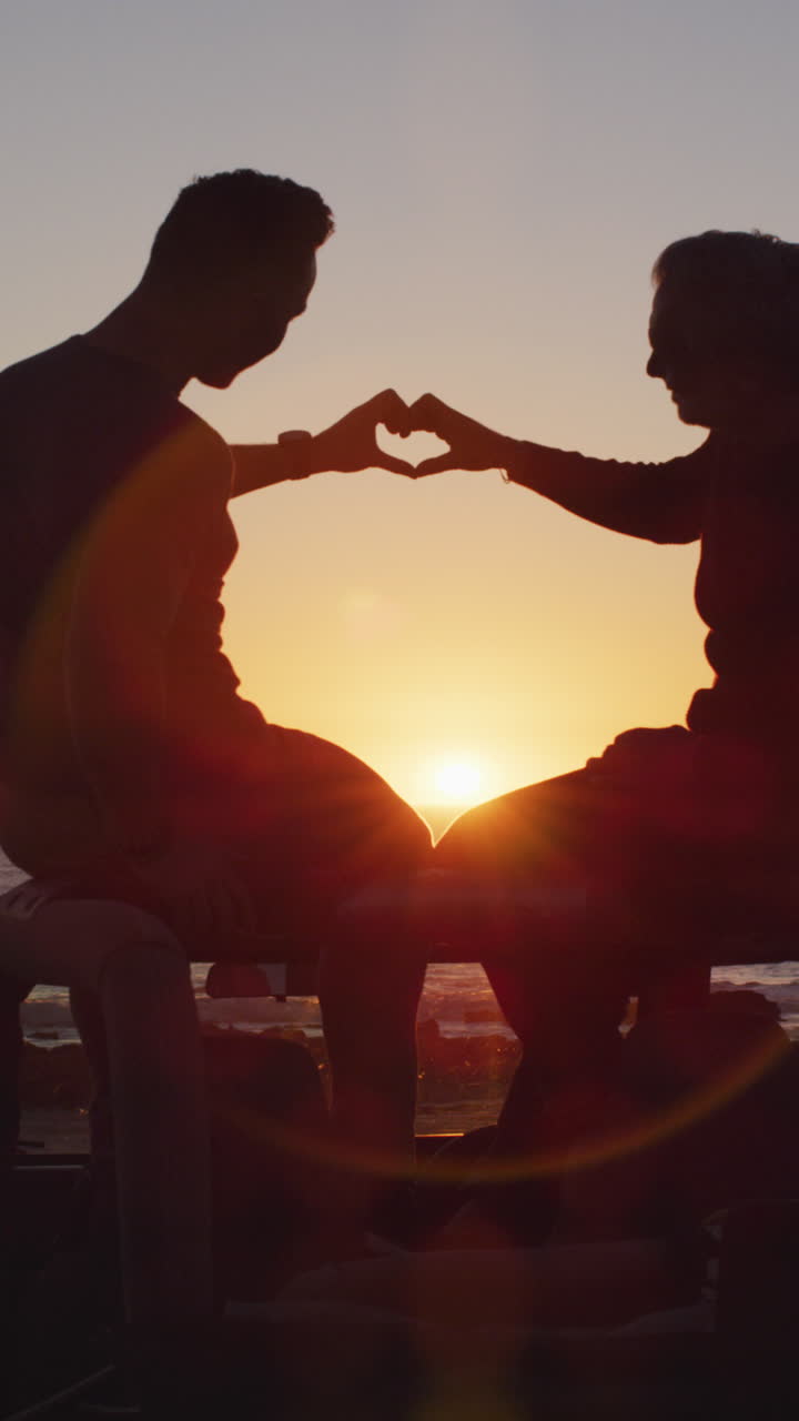 백인 동성 남성 커플이 해가 지는 해변에서 차에 앉아 손으로 심장을 만드는 수직 비디오