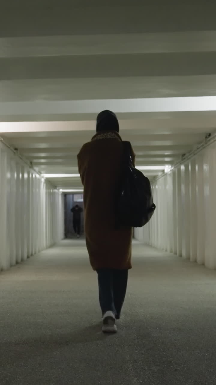 un retrato de una chica con un abrigo marrón, vaqueros, zapatos blancos y una mochila mientras camina lenta y débilmente a través de un paso subterráneo o túnel largo y débilmente iluminado