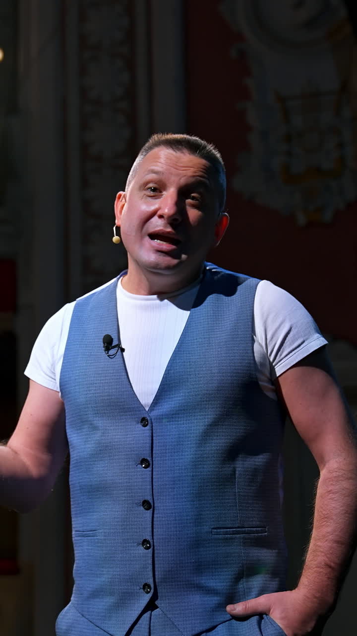 Showman entertains the audience under the light, front view. Big stage. Serious performer speaks from the stage. Hands in pockets. Vertical video