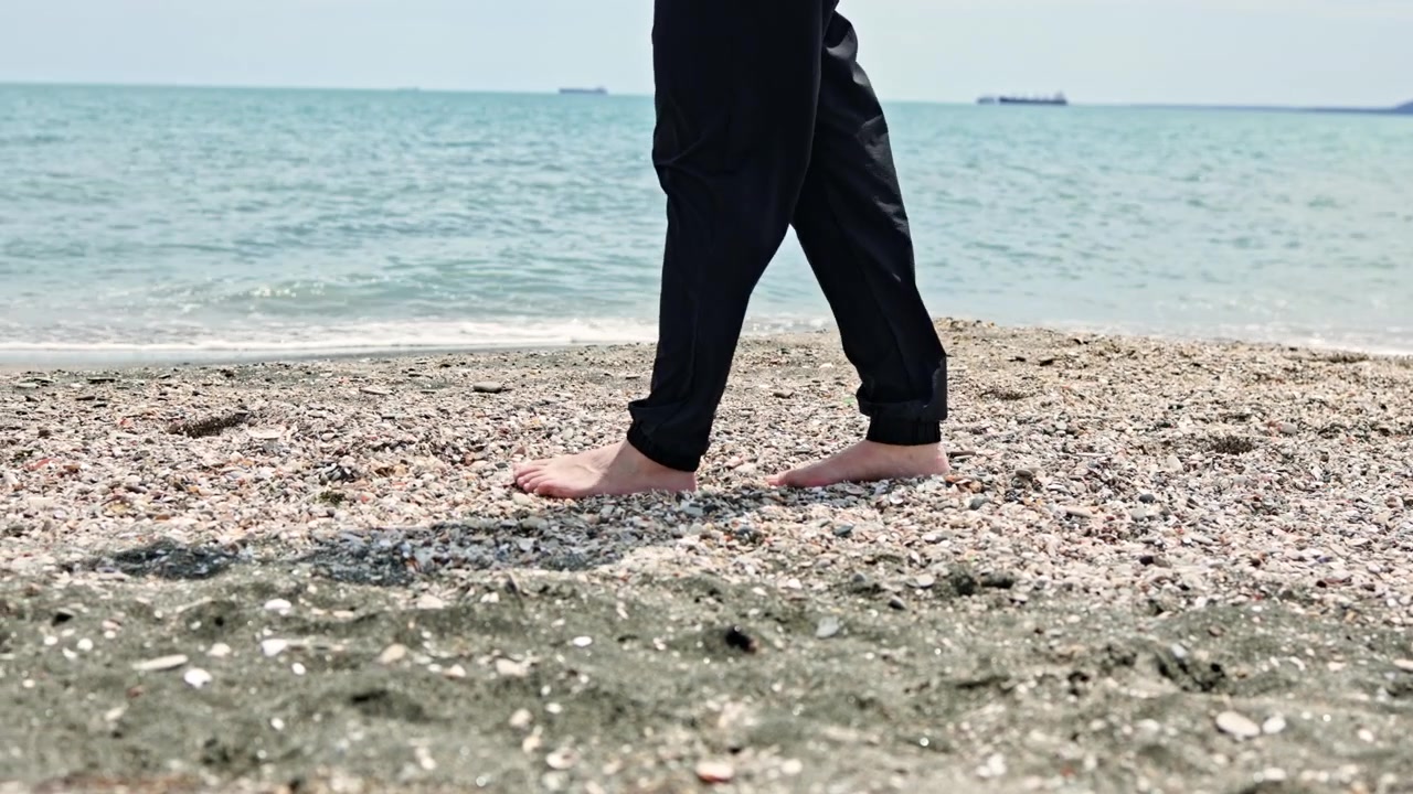 Mavi gökyüzü altında, sakin dalgalı bir sahilde çıplak ayakla duran ve uzaktaki gemilere bakan kişi.