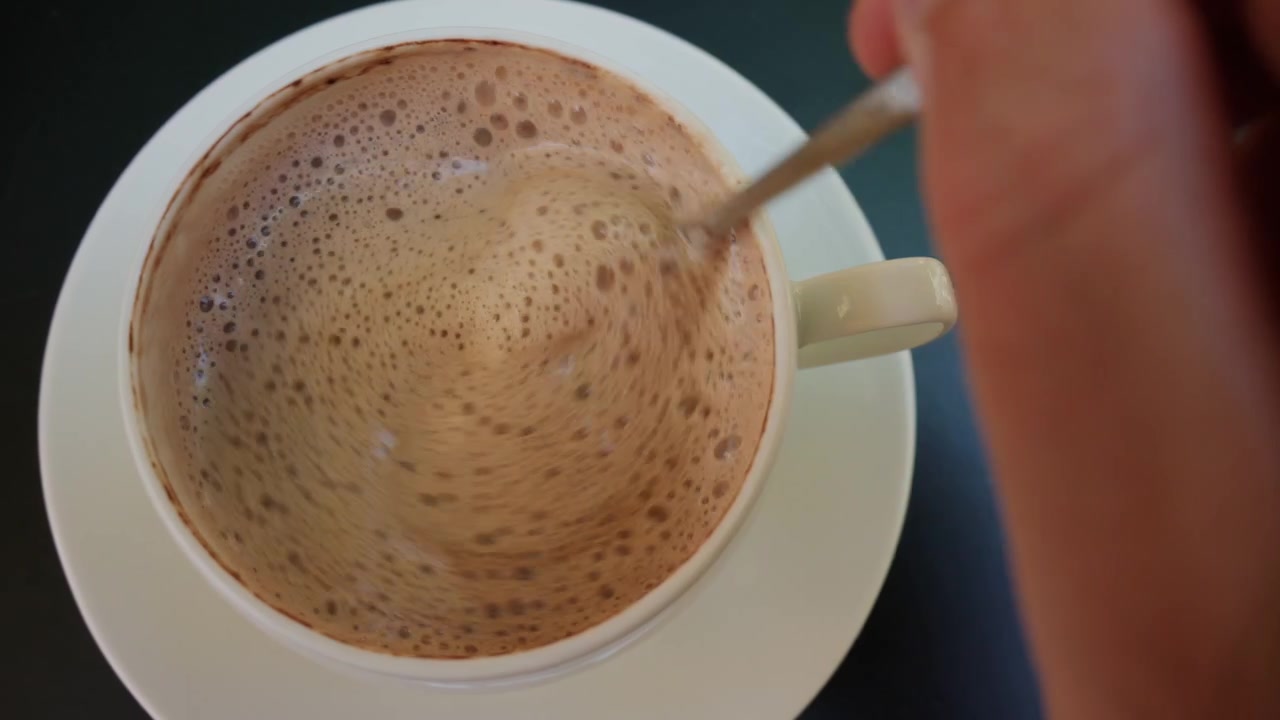 Persona che mescola un cappuccino schiumoso con un cucchiaino in una tazza bianca, creando un vortice di caffè e schiuma di latte