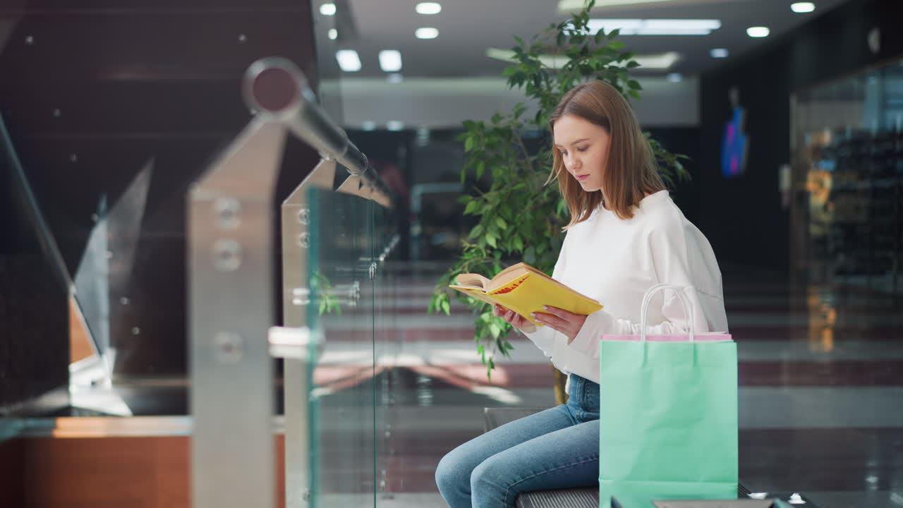 mujer leyendo un libro mientras está sentada en el centro comercial con bolsas de compras cerca, la luz natural mejora el entorno interior, la decoración moderna y las superficies reflectantes en el fondo