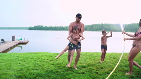 Mamá-Papá-Y-Dos-Hijos-En-El-Verano-En-El-Césped-Vertieron-Agua-De-Una-Manguera-Para-El-Césped.-Zona-De-Casa-De-Campo-Cerca-De-Un-Lago-Al-Atardecer