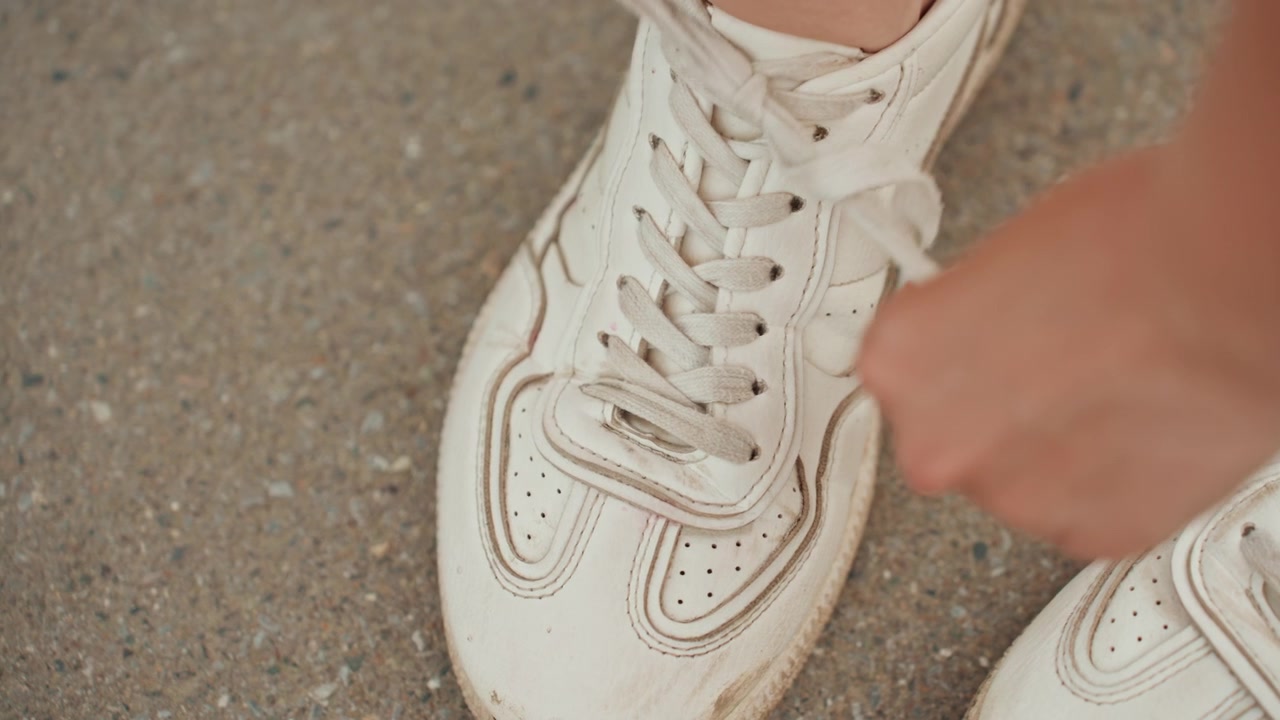 Una vista ravvicinata mostra mani che allacciano i lacci di scarpe da ginnastica bianche su una superficie di cemento ruvido. La messa a fuoco rimane stabile, catturando movimenti dettagliati mentre le dita intrecciano e tirano per fissare i lacci.