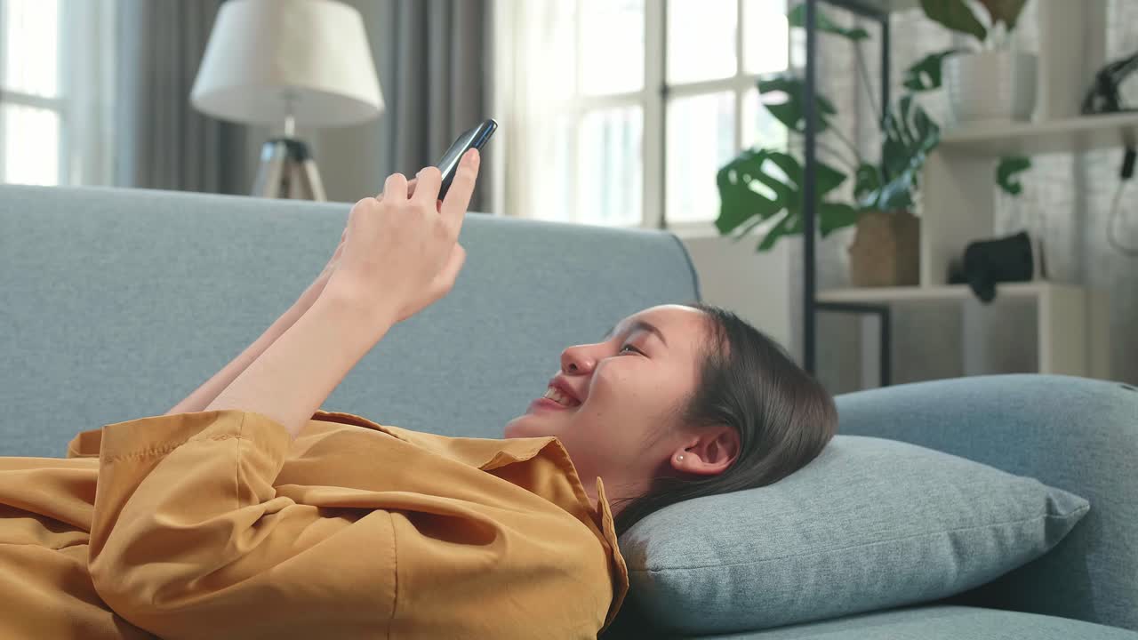 Asian Woman Lying On Couch In Living Room While Using Smartphone
