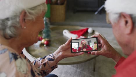 pareja caucásica mayor sonriente usando un teléfono inteligente para una videollamada de navidad con una mujer en la pantalla