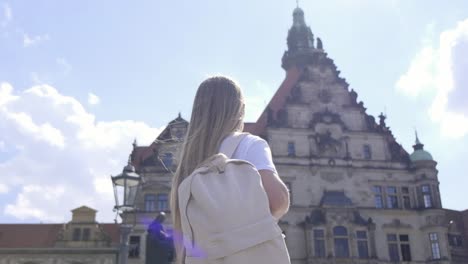 a young woman with long hair and a backpack explores the city, admiring a large building and showcasing her curiosity for architectural beauty