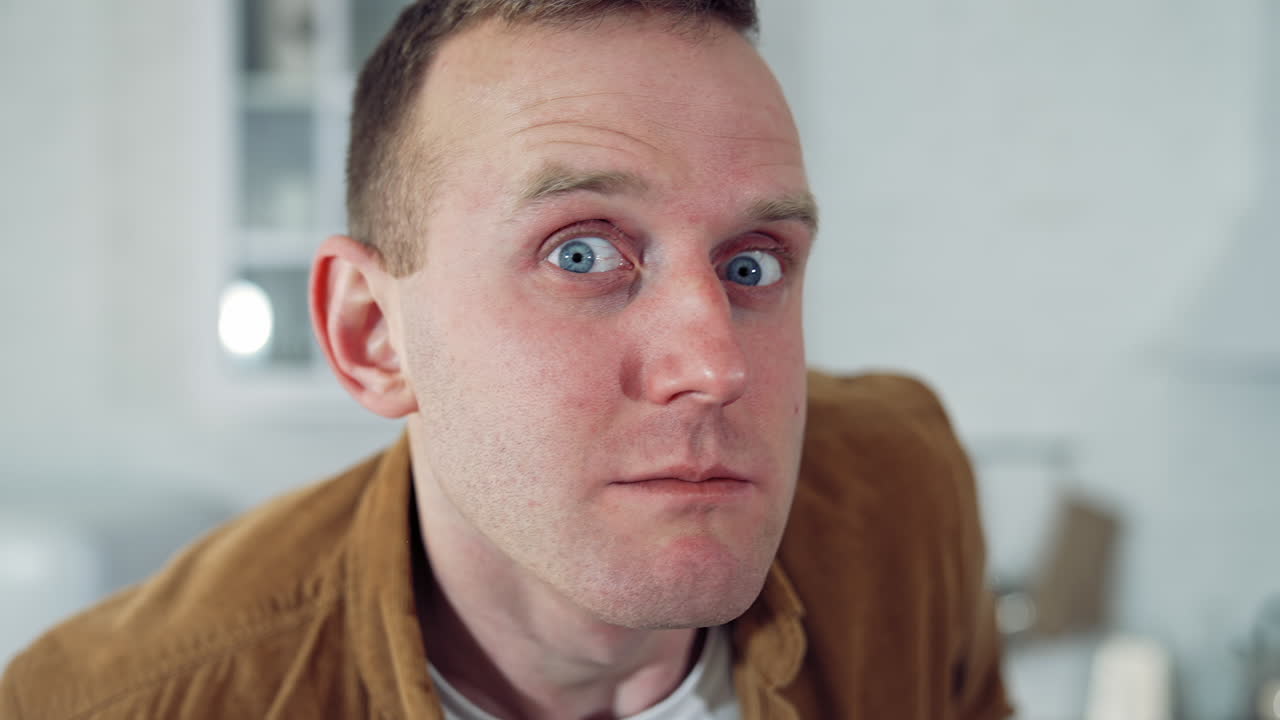 Portrait of a funny freelancer. Young man blinking eyes madly after sitting at home for a long period. Amusing man with a laptop on table in the kitchen. Lockdown concept.