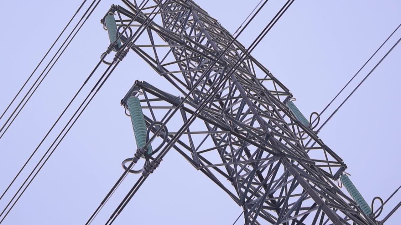 Bottom up view of a massive high voltage power pylon coated in ice and hoarfrost, with frozen suspension insulators and cables under a pale winter sky during extreme cold