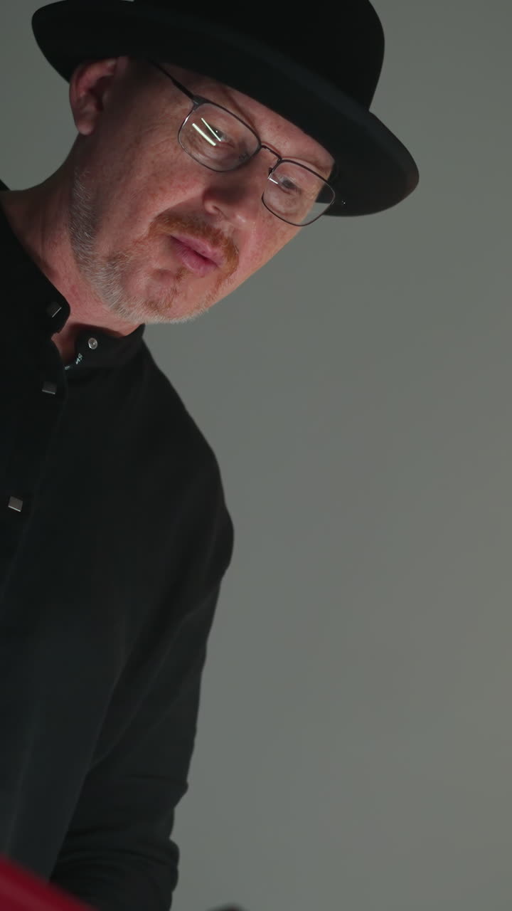 primer plano de un tecladista con un traje negro y sombrero tocando un sampler rojo, en un fondo gris