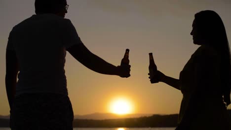 young cute latin couple toasting with beer bottles standing on a floating dock on a lake at sunset