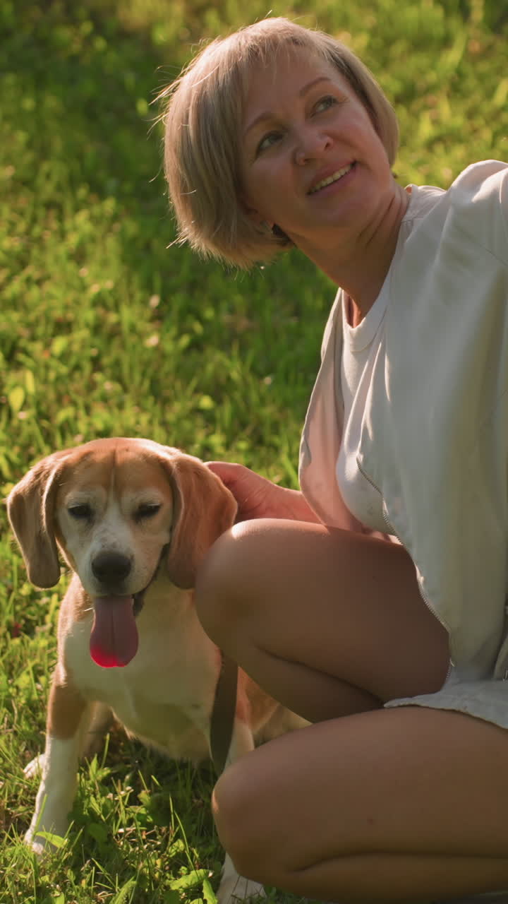 반려동물 애호가가 개와 함께 셀피를 찍고 빛 날씨 아래 울창한 녹색 정원에서 다른 무언가에 주의를 분산합니다. 배경에는 나무와 건물이 있습니다.