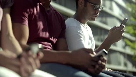 focused male friends sitting in park and using smartphones.