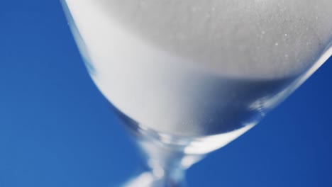 Video-of-close-up-of-hourglass-with-white-sand-pouring-with-copy-space-on-blue-background