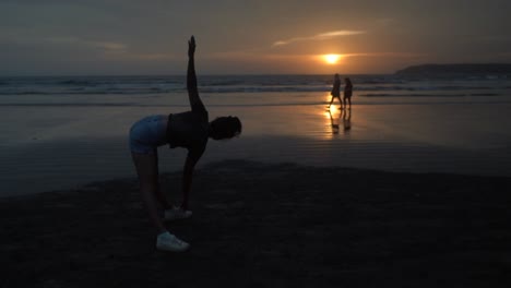 landscape view of a young woman silhouette doing stretching exercises, on a sandy beach by the ocean, at sunrise